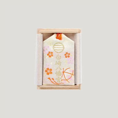 Back view of the Child Blessing Omamori from Shirasaki Hachimangu Shrine, cream embroidered charm with floral motifs inside a wooden box.