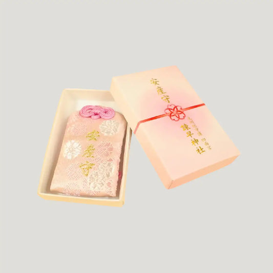 A gift set of a traditional Japanese omamori for childbirth well-being from Isahaya Shrine, featuring a pink and white floral charm and a pale pink paper gift box with a ribbon.