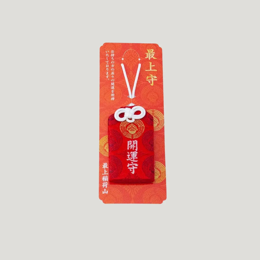 A traditional Japanese omamori for good fortune from Saijo Inari temple, with red and gold fabric and the kanji for 'good fortune', shown on its original card.