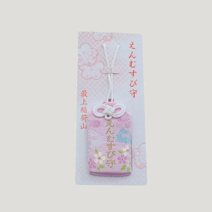 A traditional Japanese omamori for love and relationships from Saijo Inari temple, showing a light peach fabric with floral embroidery on its original card.