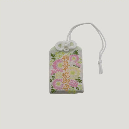 Front view of a traditional Japanese omamori for sickness recovery from Saijo Inari temple, with a white fabric and delicate floral embroidery.