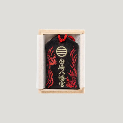Wish Fulfillment Omamori (心願成就お守り) back side – black charm with golden Shirasaki Shrine crest and red phoenix motif, presented in a wooden box